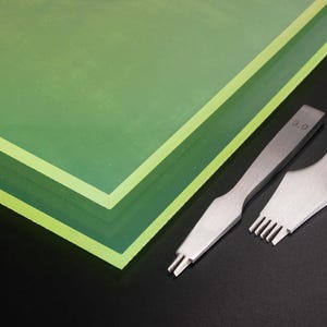 Op de afbeelding: Een stapel groene acrylplaten met een lichtere groene rand en twee zilveren leerbewerkingsgereedschappen. Eén gereedschap heeft een 3.0-markering. De gereedschappen zijn ontworpen om gelijkmatig verdeelde gaten in leer te maken om te naaien.