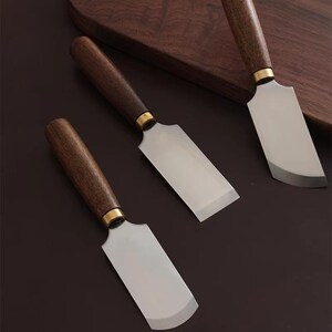 Peut inclure: Trois couteaux à fromage avec des manches en bois et des lames en acier inoxydable. Les manches sont de couleur marron foncé, avec une bande dorée où le manche rencontre la lame. Les lames ont une forme rectangulaire avec un bord incurvé.