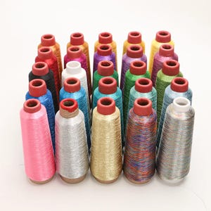 Könnte beinhalten: Eine Sammlung von fünfundzwanzig konischen Spulen aus metallischem Stickgarn, die in einem Raster angeordnet sind. Die Spulen weisen eine Vielzahl lebendiger Farben auf, darunter Pink, Silber, Gold, Blau, Grün, Violett und mehrfarbige Fäden mit glänzendem Finish.