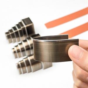 May include: A close-up of metal leather working tools. The main tool is a curved, dark metal piece with a silver edge, held by a hand. Three stepped metal tools are in the background, along with two orange strips.
