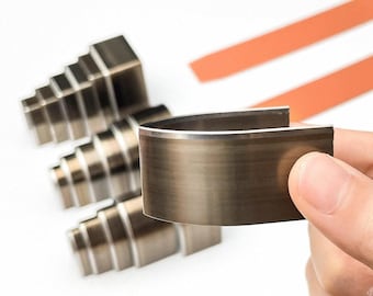 Może przedstawiać: Zbliżenie metalowych narzędzi do obróbki skóry. Głównym narzędziem jest zakrzywiony, ciemny metalowy element ze srebrną krawędzią, trzymany w dłoni. Trzy schodkowe metalowe narzędzia znajdują się w tle, wraz z dwoma pomarańczowymi paskami.