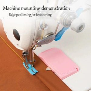 May include: A white sewing machine with a blue presser foot stitching a brown fabric. A pink plastic edge guide with a ruler is positioned on the machine bed. The text reads: Machine mounting demonstration. Edge positioning for topstitching.