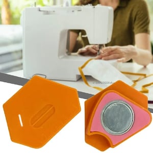 May include: Two orange plastic sewing seam guides with a pentagonal shape. One guide shows a moulded slot and a ruler edge. The other guide features a circular metal magnet insert surrounded by a pink border, designed for alignment on a sewing machine.