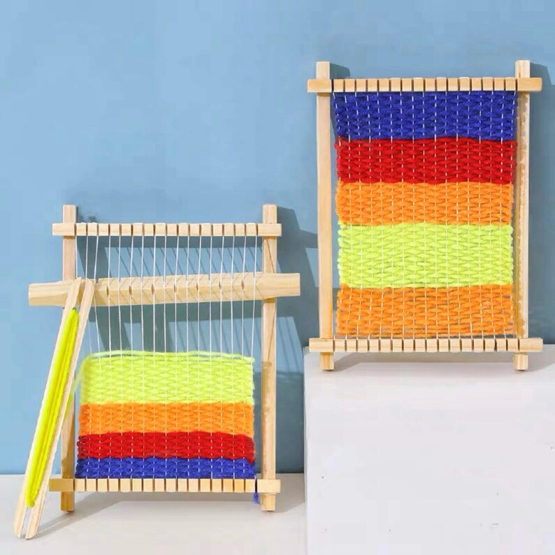 May include: Two wooden weaving looms with colorful yarn. The yarn is woven in horizontal stripes of blue, red, orange, yellow, and orange. The looms are made of light-colored wood and have a rectangular frame.