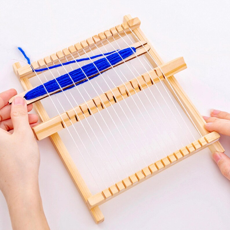 May include: A small wooden weaving loom with white warp threads and blue yarn being woven through. The loom is rectangular with notches along the top and bottom edges. A hand holds the shuttle with the blue yarn.
