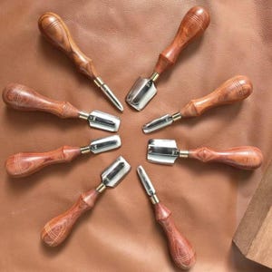 May include: A collection of seven leathercrafting tools arranged in a circular pattern. Each tool features a polished wooden handle with a distinct grain pattern and a metallic ferrule connecting it to a sharp, silver-colored steel blade.