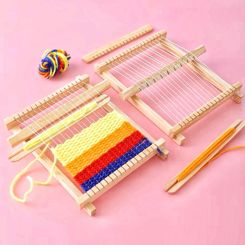 May include: Wooden weaving looms with white string and colorful yarn. One loom has a partially woven piece with yellow, orange, red, and blue yarn. A ball of multi-colored yarn and a weaving needle are also present.