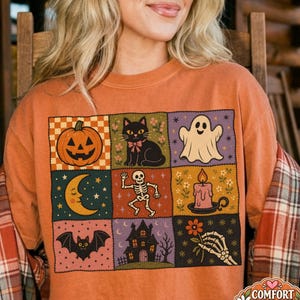 May include: A burnt orange t-shirt featuring a grid of nine square illustrations including a jack-o'-lantern, a black cat, a ghost, a crescent moon, a skeleton, a candle, a bat, a haunted house, and a skeletal hand holding a flower.