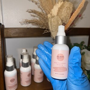 May include: A clear glass bottle with a silver pump top and a white label. The label says "SKIN ORGANIC" and "BIKINI BODY SERUM" with a pink and white design. The bottle is labeled "NET WT 2 FL OZ (59 ML)".