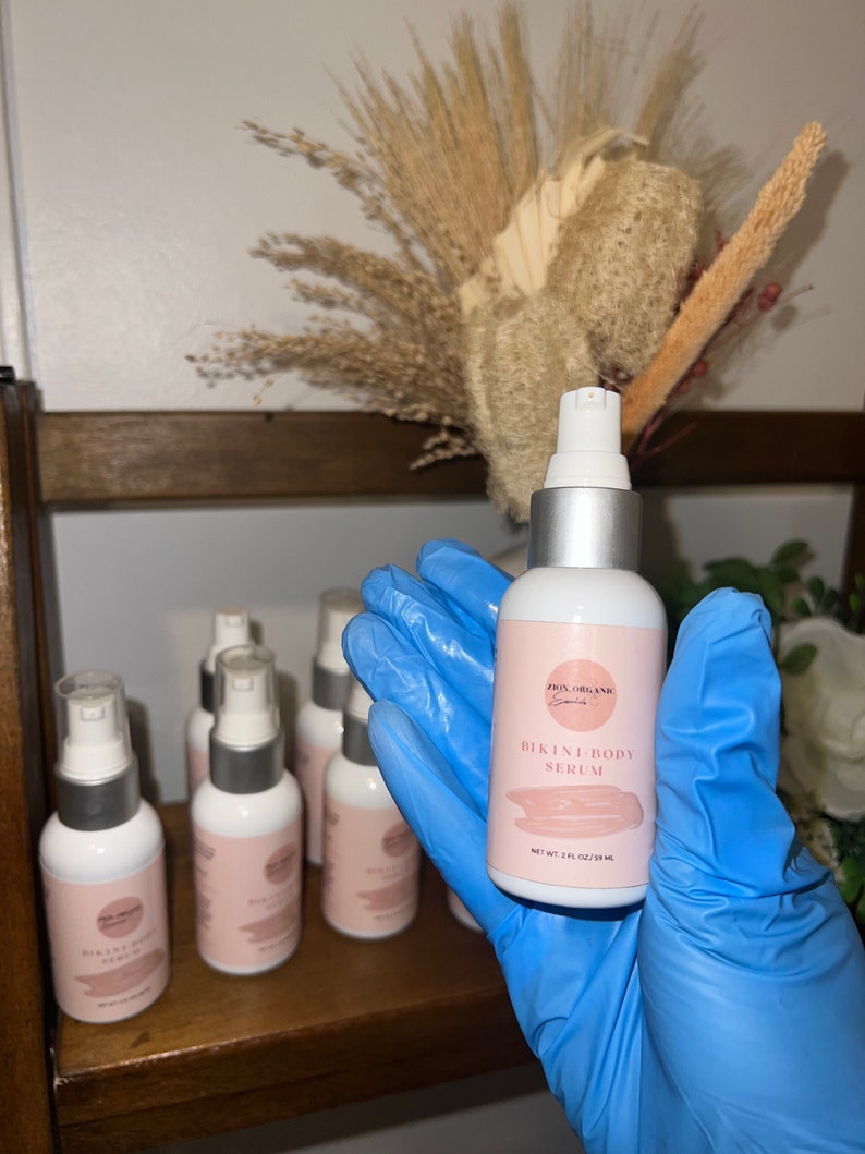 May include: A clear glass bottle with a silver pump top and a white label. The label says "SKIN ORGANIC" and "BIKINI BODY SERUM" with a pink and white design. The bottle is labeled "NET WT 2 FL OZ (59 ML)".