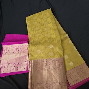 May include: A folded chartreuse saree with a gold floral pattern and a gold and pink border. A matching pink fabric with a gold floral border is folded next to the saree. The saree is displayed on a black surface.