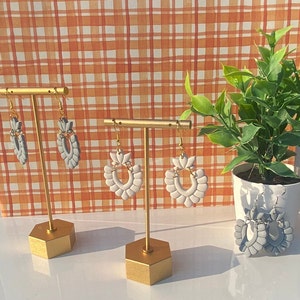 May include: Three pairs of gray and white ceramic earrings with a scalloped design. The earrings are displayed on gold metal earring stands against a checkered background.