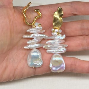 May include: A pair of gold-toned earrings featuring a unique design. Each earring has a gold top, a cluster of small pearls, a stack of white, irregular-shaped pearls, and a large, iridescent pearl at the bottom. The earrings are held in a hand.