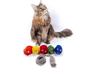 May include: A Maine Coon cat sits and looks up at a collection of colorful cat toys. The toys include felted balls in red, yellow, green, and blue, each with a smiling face, and a gray felt mouse and a gray felt ball.