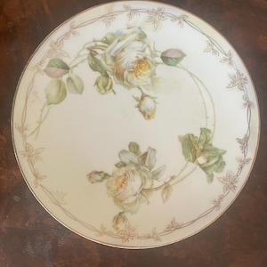 May include: A round porcelain plate with a cream-colored base featuring two clusters of pale yellow and white roses with green leaves. The rim is decorated with a delicate, thin gold-toned filigree pattern of vines and leaves encircling the edge.