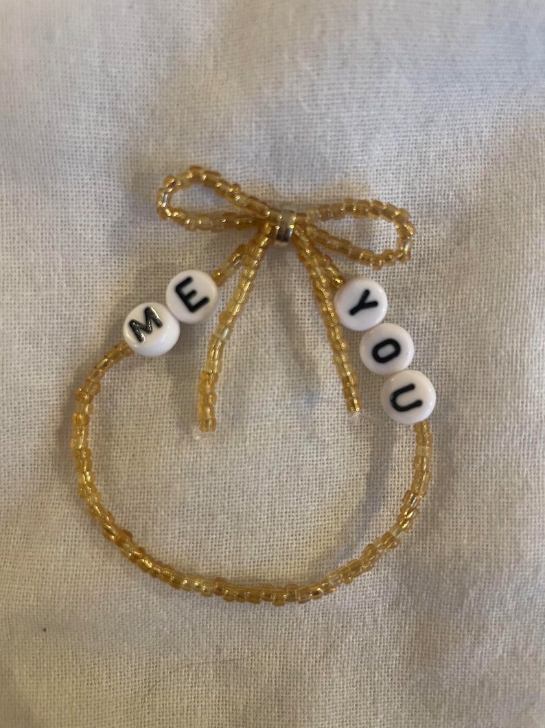 Puede incluir: Un brazalete de cuentas doradas con un lazo en la parte superior y cuentas de letras blancas que deletrean "ME YOU".
