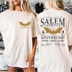 May include: Cream-colored t-shirt with a gold bat graphic and the word "CUSTOM" on the front. The back of the shirt features a gold bat design and the words "SALEM MEDICINES APOTHECARY" with additional text and design elements.