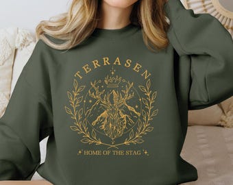 May include: A dark green sweatshirt with a gold graphic design. The design features the word "TERRASEN" in an arch, a stag head with a crown, mountains, and the text "HOME OF THE STAG". The design is surrounded by a laurel wreath.