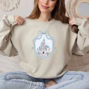 A woman is sitting on a bed, wearing a beige sweater with a castle design on it.
