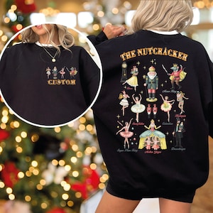 May include: Black sweatshirt with a white collar featuring a "CUSTOM" design on the front and a "THE NUTCRACKER" graphic on the back. The back graphic includes illustrations of characters from the ballet, such as the Sugar Plum Fairy and the Mouse King.