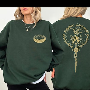 May include: Dark green sweatshirt with a gold ring design on the front and a gold sword and inscription design on the back. The sweatshirt has a crew neck and long sleeves. The design is inspired by a fantasy theme.