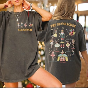 May include: Dark gray t-shirt featuring a custom design with ballet figures and the text "CUSTOM" on the front. The back of the shirt displays "THE NUTCRACKER" and various characters from the ballet. A comfortable, casual style.