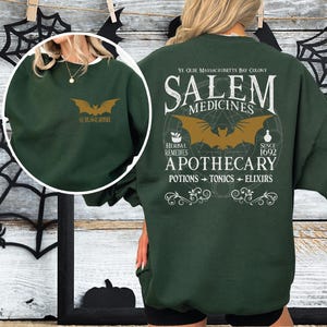 May include: Dark green sweatshirt with a gold bat graphic and the word "CUSTOM" on the front. The back features a Salem Apothecary design with a bat, text, and the words "Herbal Remedies" and "Since 1692".