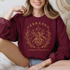 a woman sitting on a bed wearing a maroon sweatshirt