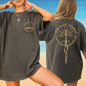 A woman wearing a gray t-shirt with a tree design on the back, posing on a beach with a blue sky and ocean in the background.