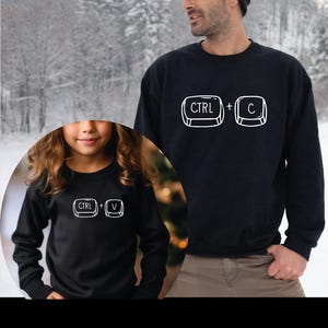 May include: Black sweatshirts with white graphic keyboard symbols. One sweatshirt displays "CTRL + C", and the other displays "CTRL + V". The sweatshirts are worn by a person and a child, with a snowy background.