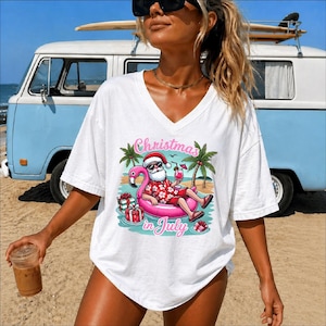 May include: A white V-neck t-shirt featuring a graphic print of Santa Claus wearing sunglasses and a red floral shirt, relaxing in a pink flamingo pool float. The design includes palm trees, wrapped gifts, and the text Christmas in July.
