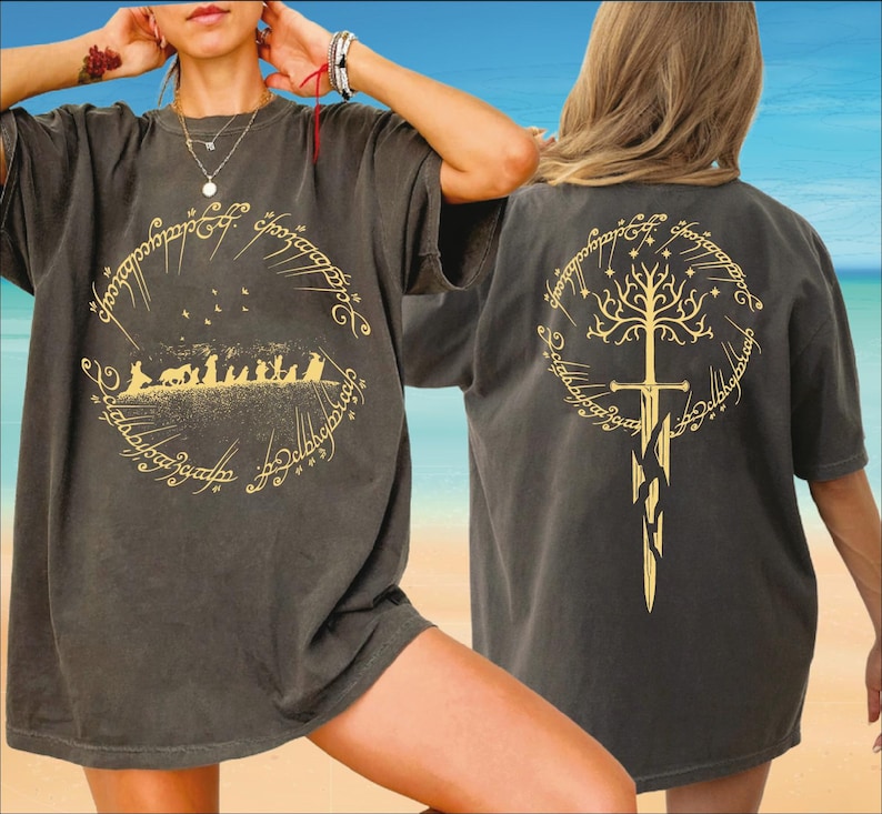 A person wearing a black t-shirt with a graphic design featuring a tree and a sword, standing on a beach with a clear blue sky in the background.