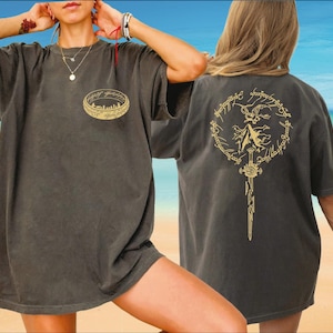 May include: Dark gray t-shirt featuring a gold ring design on the front and a circular graphic with a sword on the back. The front design includes a ring with a city silhouette. The back design has text around the circular graphic.