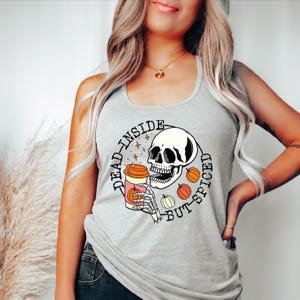 May include: A light gray tank top featuring a graphic print of a skull holding a coffee cup with a pumpkin design. The text "DEAD-INSIDE BUT-SPICED" is printed in a circular pattern around the skull, accompanied by small orange pumpkin illustrations.