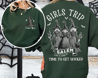May include: Dark green sweatshirt with a white collar. The front features a witch hat graphic with a crescent moon and stars, and the text "Custom Name." The back displays the words "GIRLS TRIP" above a graphic of three witches, with "SALEM" and "TIME TO GET WICKED" below.