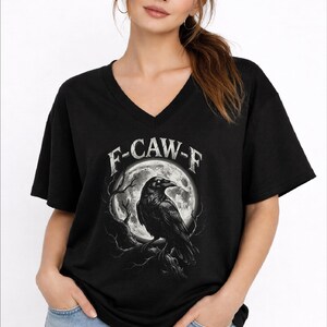 May include: A black V-neck t-shirt featuring a graphic print of a raven perched on a tree branch in front of a large, detailed full moon. The text F-CAW-F is printed in a distressed white font in an arc above the moon and the raven illustration.