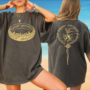 A woman wearing a black t-shirt with a graphic design on the front and a back design on the back, posing on a beach with a clear blue sky in the background.