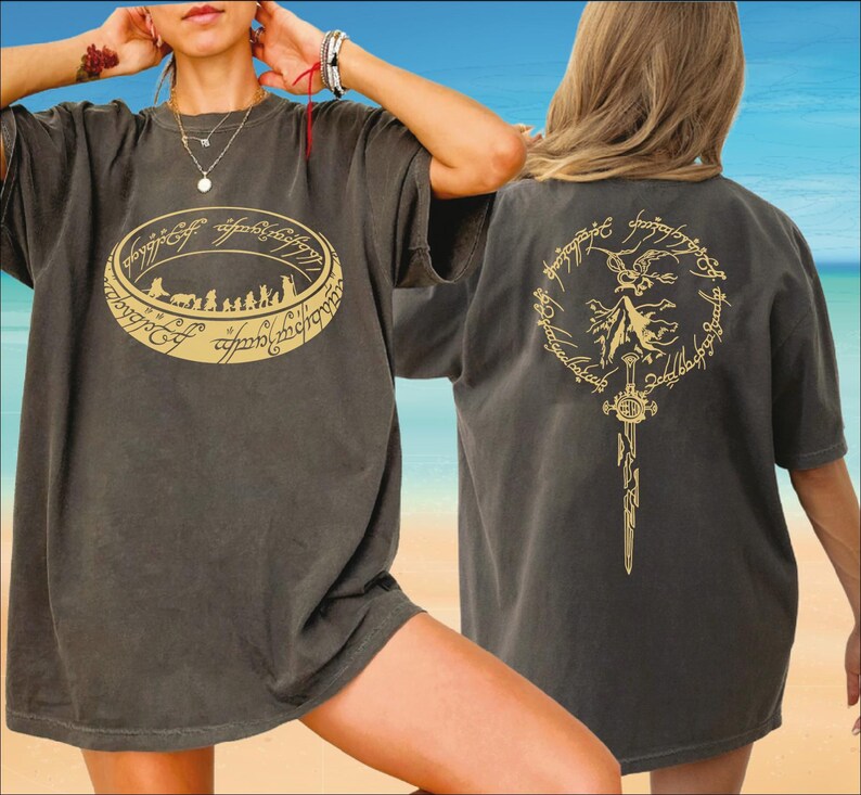 A woman wearing a black t-shirt with a graphic design on the front and a back design on the back, posing on a beach with a clear blue sky in the background.