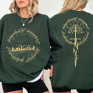 May include: Dark green sweatshirt featuring a gold design from a fantasy novel. The front displays a circular design with figures and text. The back has a sword and tree design within a circular frame. The sweatshirt is a long-sleeved, crew neck style.