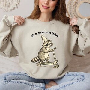 May include: Beige sweatshirt with the text "off to commit mom foolery." The graphic features a raccoon wearing a cone hat and riding a scooter. The raccoon is in shades of brown and gray.