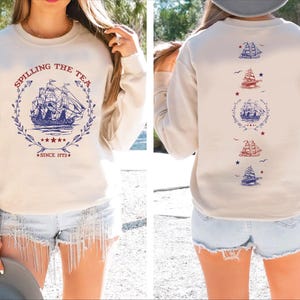 May include: Beige sweatshirt with a navy blue ship graphic inside a wreath, the text "Spilling the Tea Since 1773" is printed on the front. The back features additional ship graphics in red and blue.