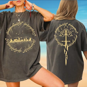 May include: Dark gray t-shirt featuring a gold design from a fantasy novel. The front displays a circular design with figures and text. The back shows a tree and sword design within a circle. The shirt is worn by a person.