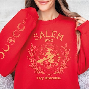 A woman wearing a red sweater with a design featuring a witch on a broomstick and the text "SALEM 1692 They Missed One" printed on it.
