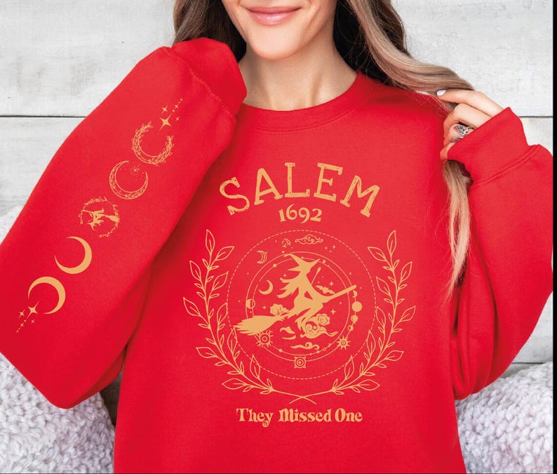 A woman wearing a red sweater with a design featuring a witch on a broomstick and the text "SALEM 1692 They Missed One" printed on it.