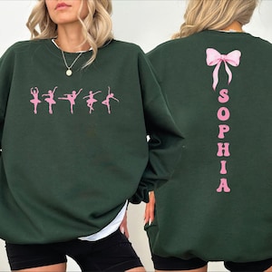 May include: Dark green sweatshirt featuring pink ballet dancer silhouettes across the front. The back of the sweatshirt displays the name "SOPHIA" in pink, with a pink bow above. The sweatshirt has long sleeves and a white trim at the neckline.