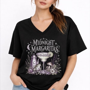 May include: A black V-neck t-shirt featuring a graphic print with the text "MIDNIGHT MARGARITAS" in a stylized font. The design includes a margarita glass, a metal pitcher, a frog, a snail, mushrooms, and floral elements in purple tones.