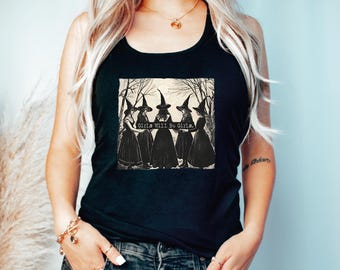 May include: A black tank top featuring a graphic print of five figures wearing pointed hats and long dresses standing in a circle among trees. The text Girls Will Be Girls is printed in white across the center of the graphic illustration.