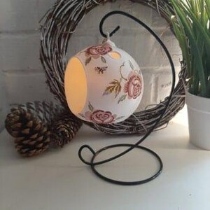 May include: White ceramic candle holder with a floral design and a bee. The holder is hanging from a black metal stand.