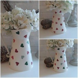 Op de afbeelding: Witte keramische kan met een patroon van groene en rode harten. De kan heeft een brede opening en een handvat.