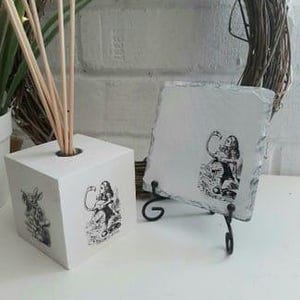 May include: White ceramic diffuser with a black and white image of Alice in Wonderland. The diffuser has a square shape and a hole in the top for reeds. The image is also on a square slate coaster with a black metal stand.